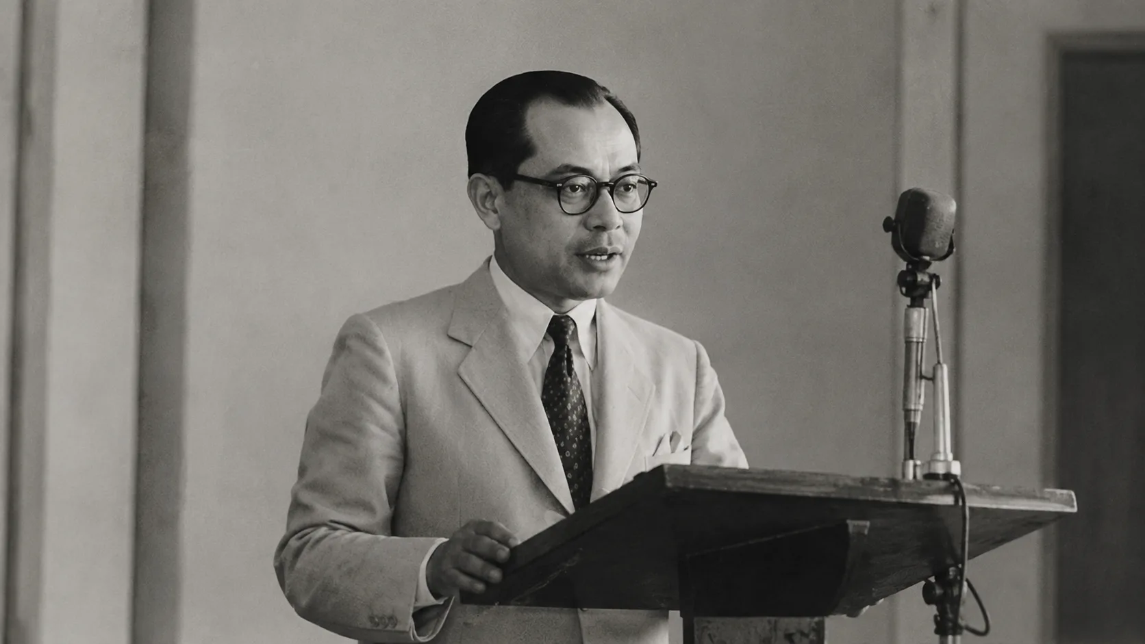 Foto hitam-putih bergaya arsip seorang pria mengenakan setelan jas berbicara di depan mikroskop podium tua, menggambarkan suasana pidato politik luar negeri era 1948.